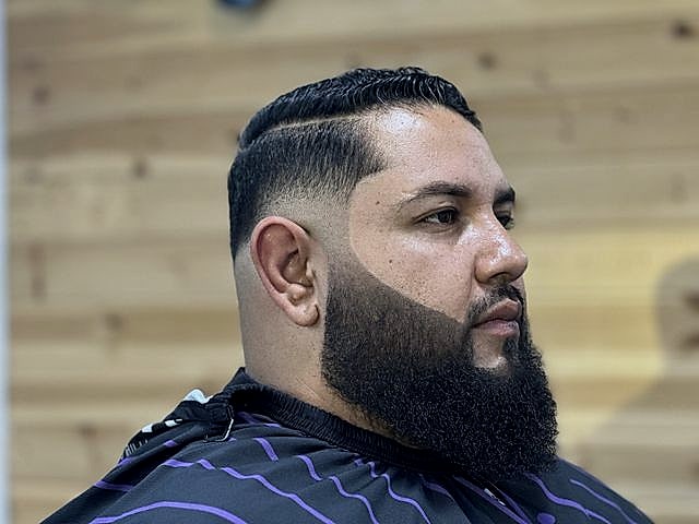 Perfil de un cliente con fade y barba larga, frente a la pared de pino.